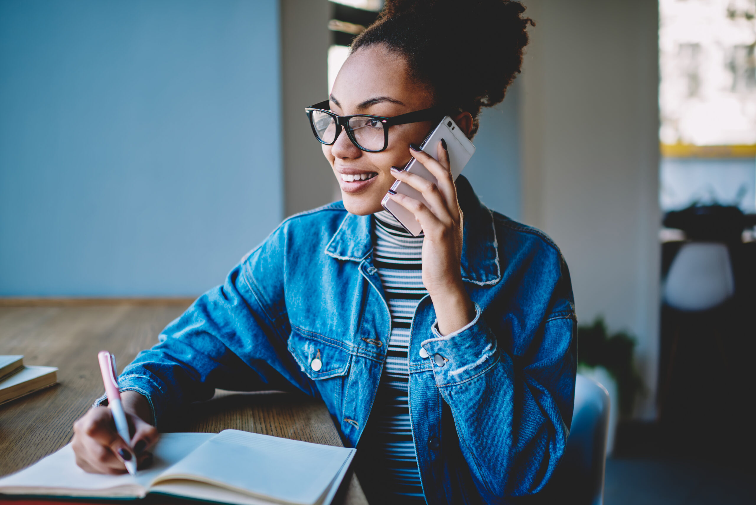 Positive female teenager in trendy spectacles for vision correction calling to friend for discussing time of meeting in cafeteria, happy hipster girl learning indoors and using mobile app for talking